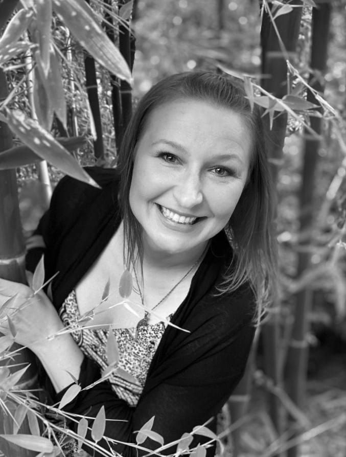 A woman is smiling in a black and white photo while standing in a forest.