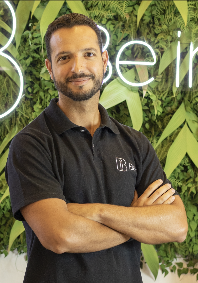 Um homem com barba está de pé com os braços cruzados em frente a uma parede verde.