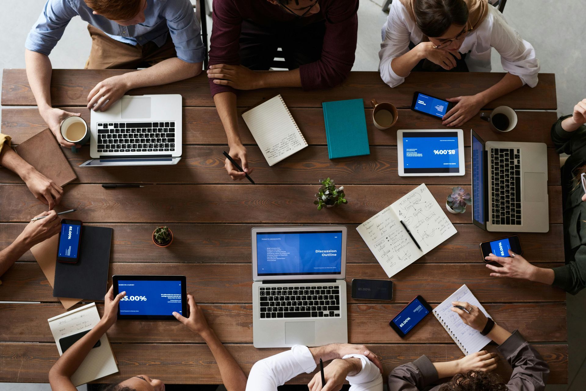 Uma mesa compartilhada com vários funcionários trabalhando em seus laptops, cadernos e celulares.