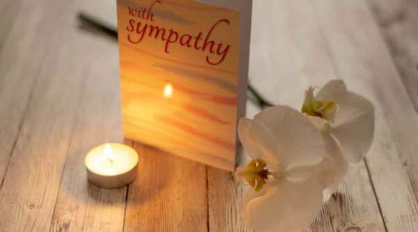 A sympathy card is sitting on a wooden table next to a candle and flowers.