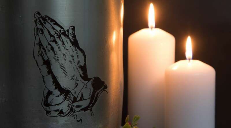 A urn with praying hands on it is next to two lit candles.