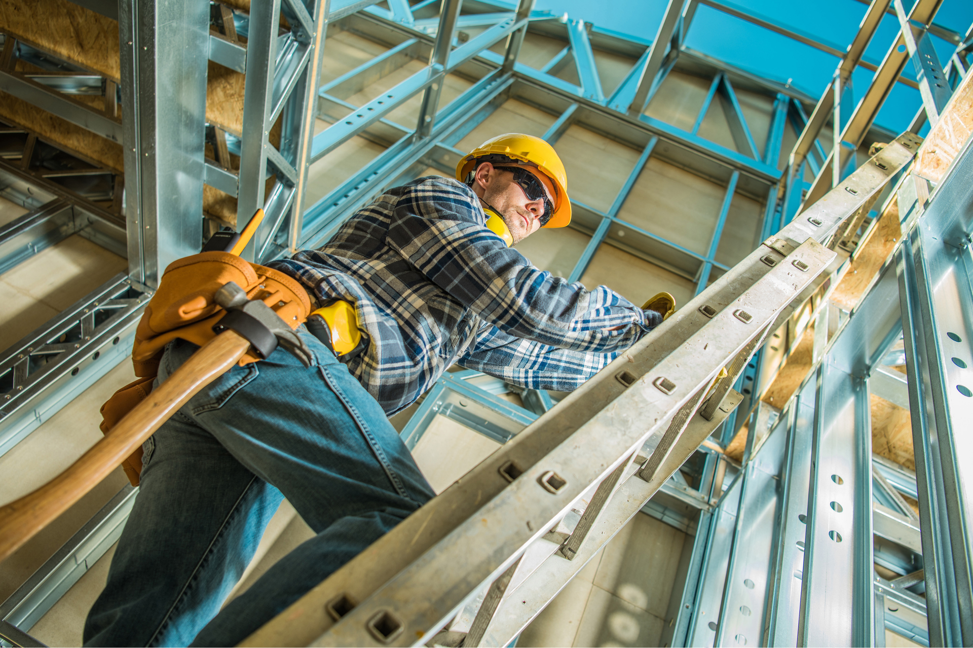 Construction worker on a ladder, working on metal framing, wearing hard hat and tool belt.