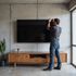 Man mounts a large, black TV on a concrete wall above a wooden entertainment center in a modern living room.