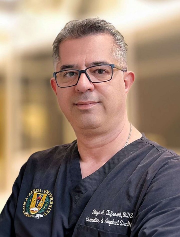 A man wearing glasses and a scrub top is standing in front of a blurry background.