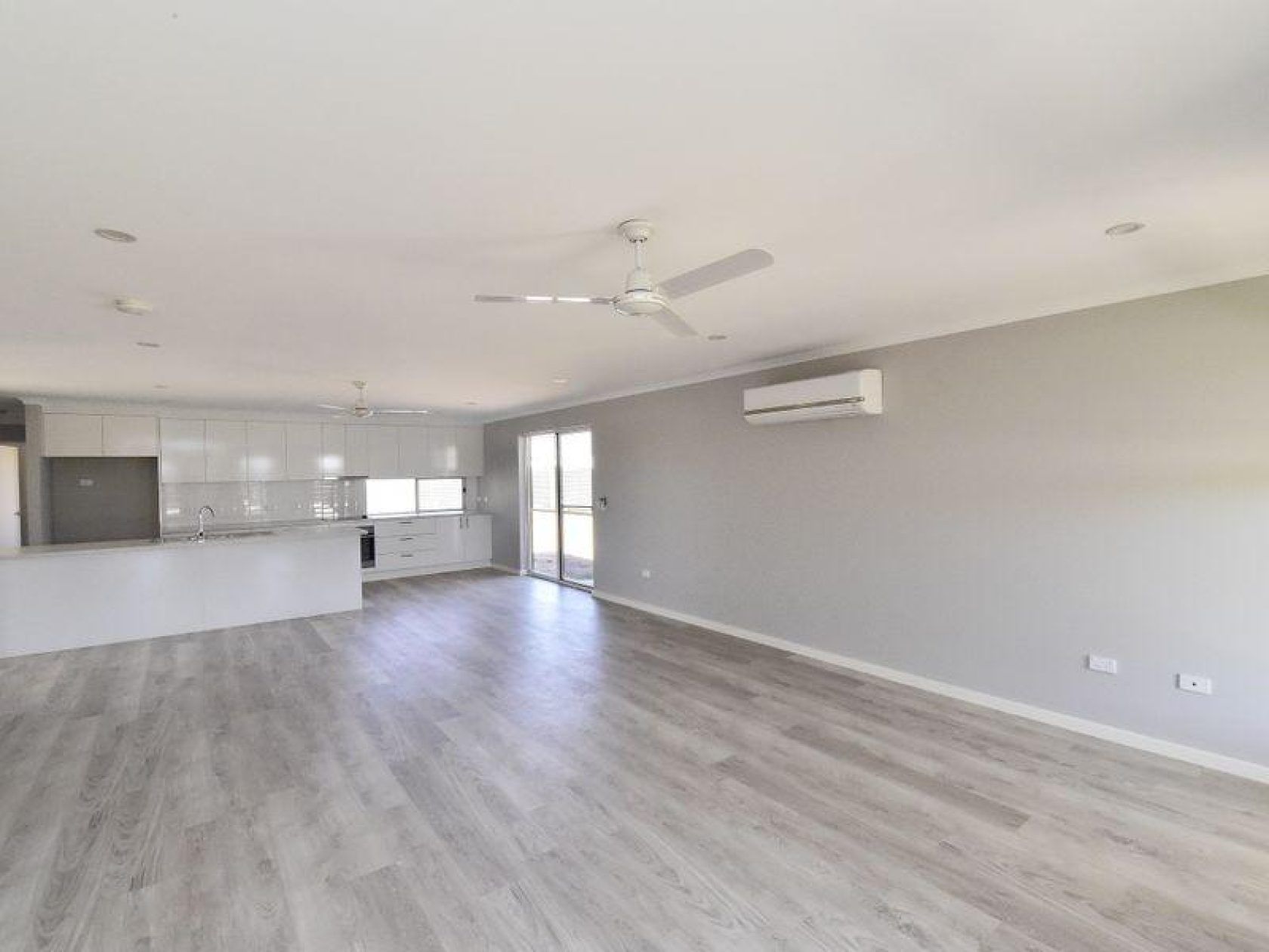 Empty, bright living area with white kitchen, gray walls, and light wood-look flooring.