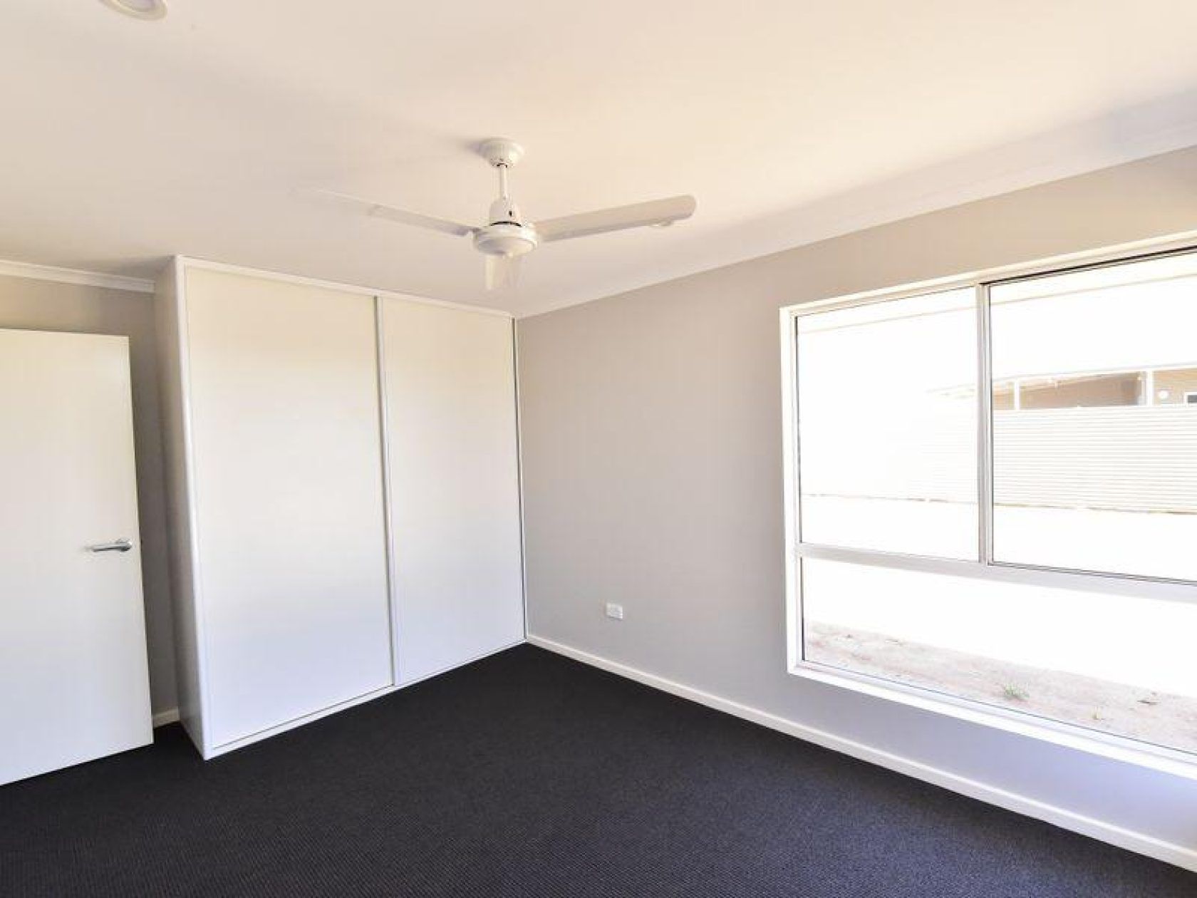 Empty bedroom with gray walls, large window, closet, dark carpet, and ceiling fan.