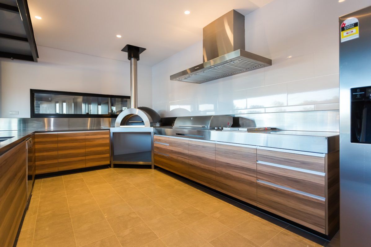 Outdoor kitchen with pizza oven, grill, stainless steel appliances, wooden cabinetry, tile floor.