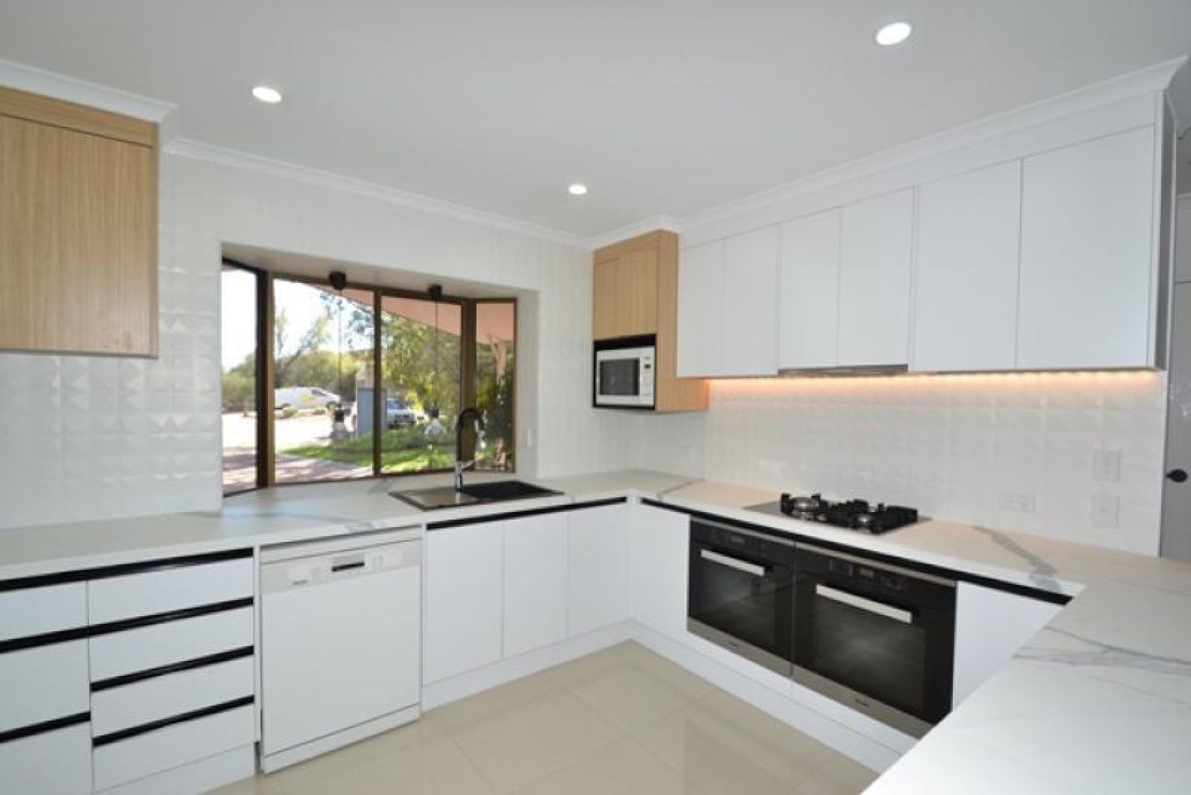 Modern white kitchen with light wood accents, stainless steel appliances, and a window overlooking a yard.