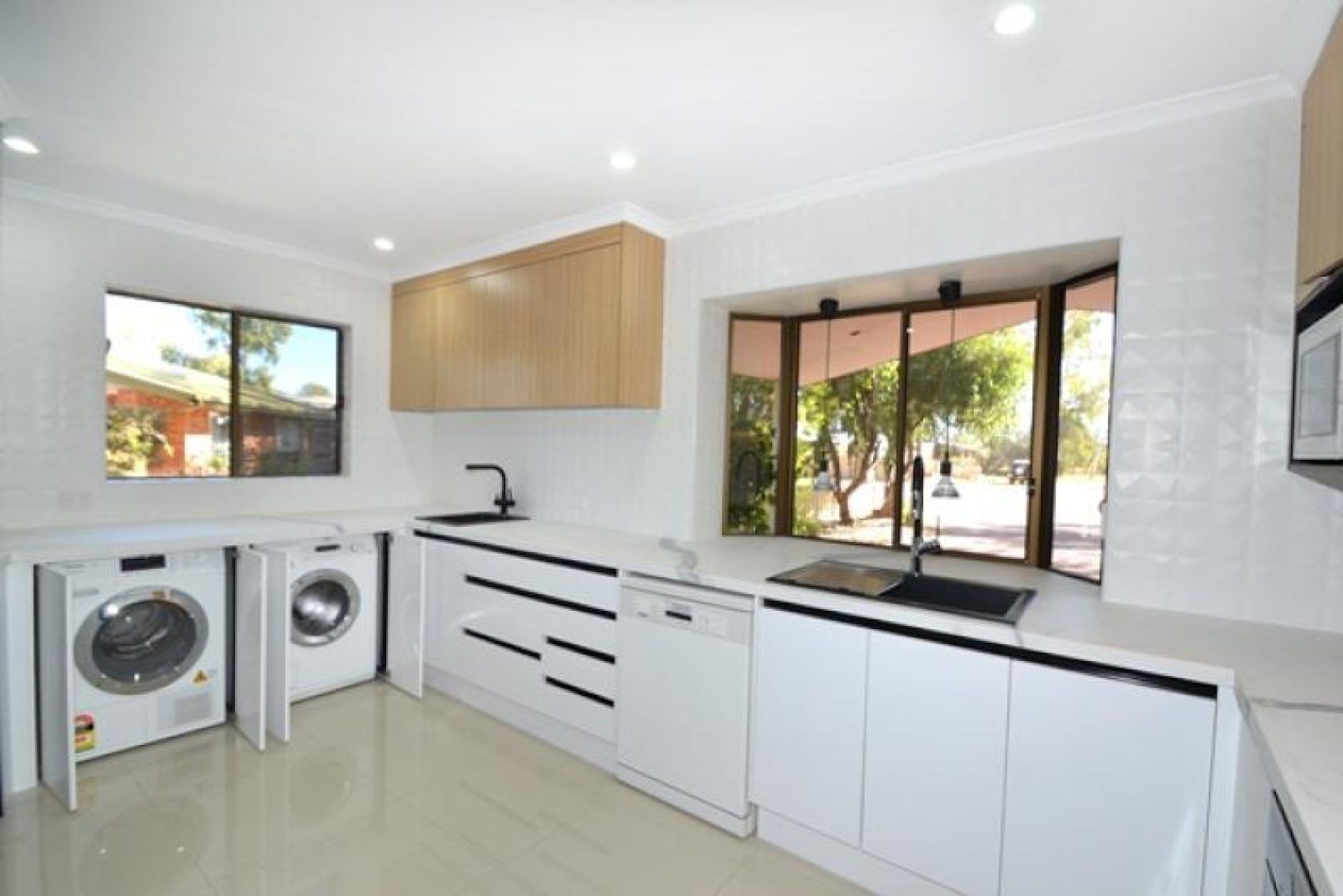 Modern white kitchen with appliances, light wood cabinets, and large window overlooking a backyard.