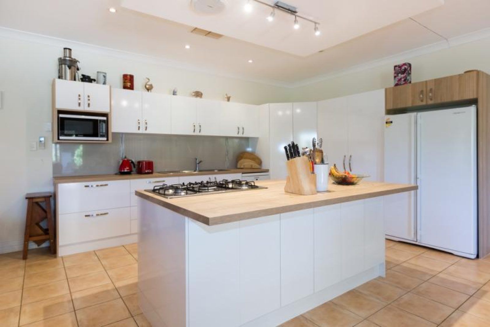 White kitchen with island, stovetop, and appliances, light wood countertops, neutral tile floor.
