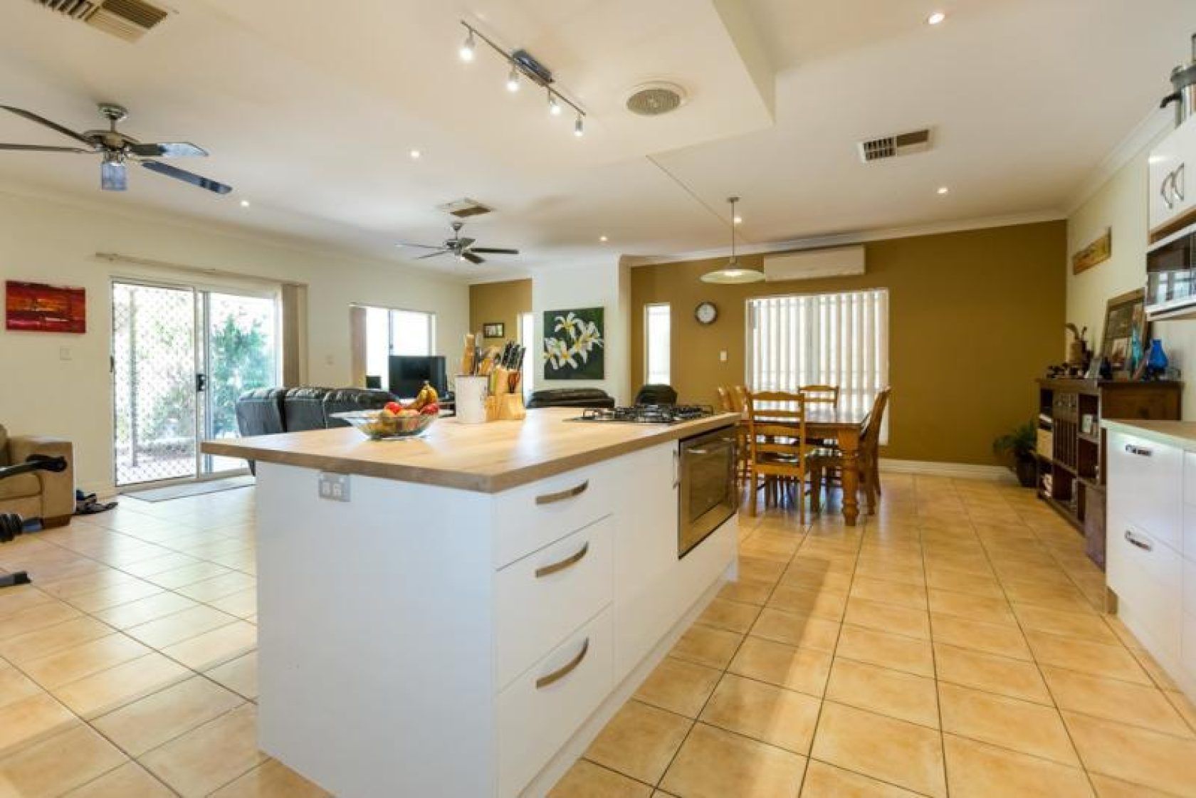 Open-concept kitchen with island, dining area, and living room. White cabinetry, wooden table, and tan tile flooring.