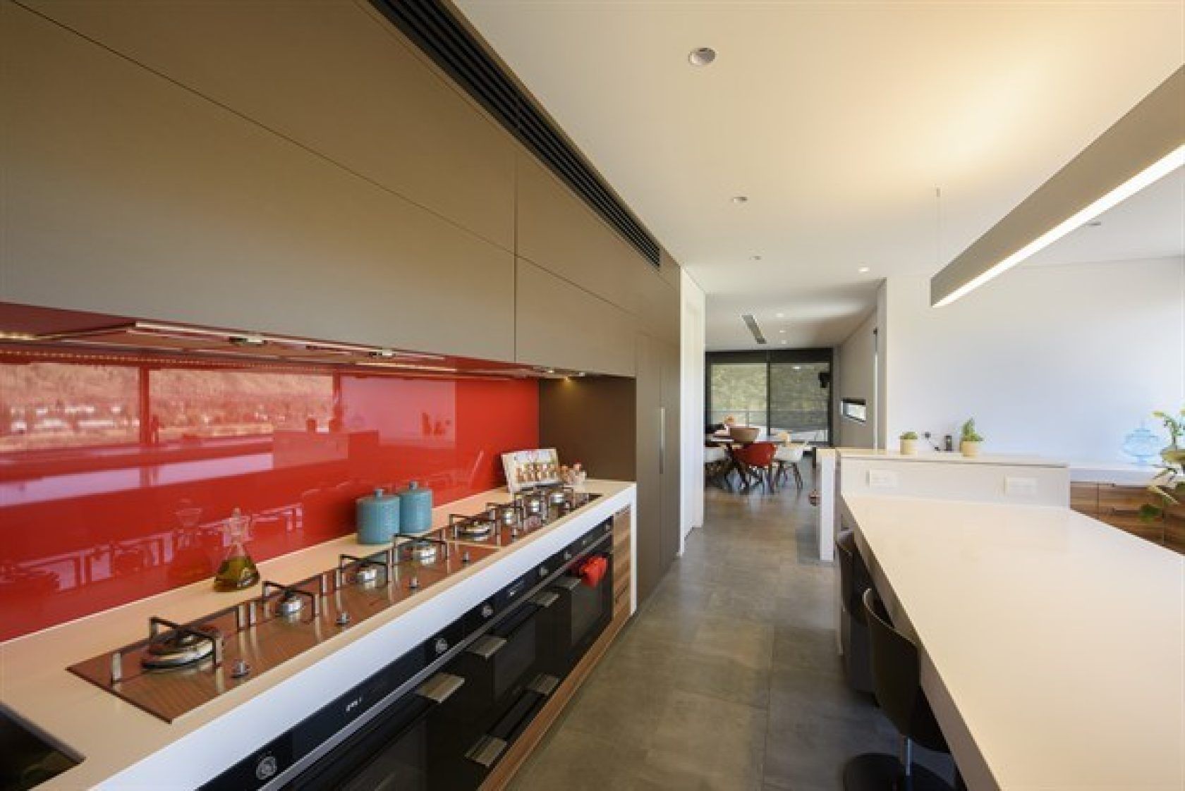 Modern kitchen with red backsplash, cooktop, ovens, and a long white countertop island.
