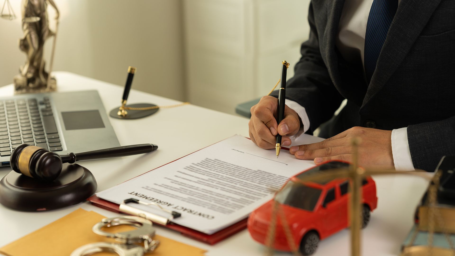 A car accident lawyer signing legal papers at a desk with a laptop, gavel, handcuffs, and toy car