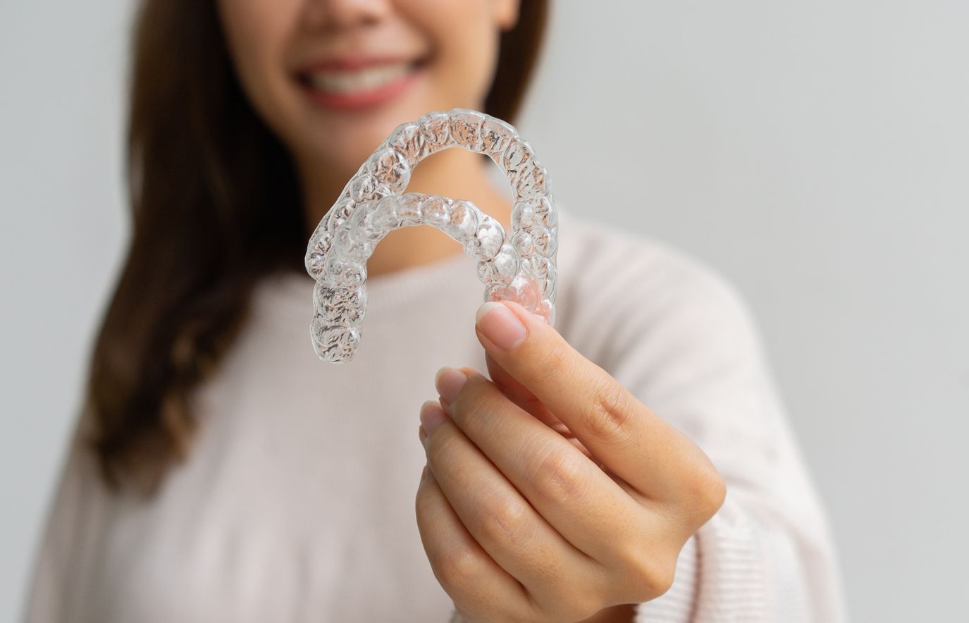 A smiling person holds a transparent dental aligner tray toward the camera against a plain light-gray background.