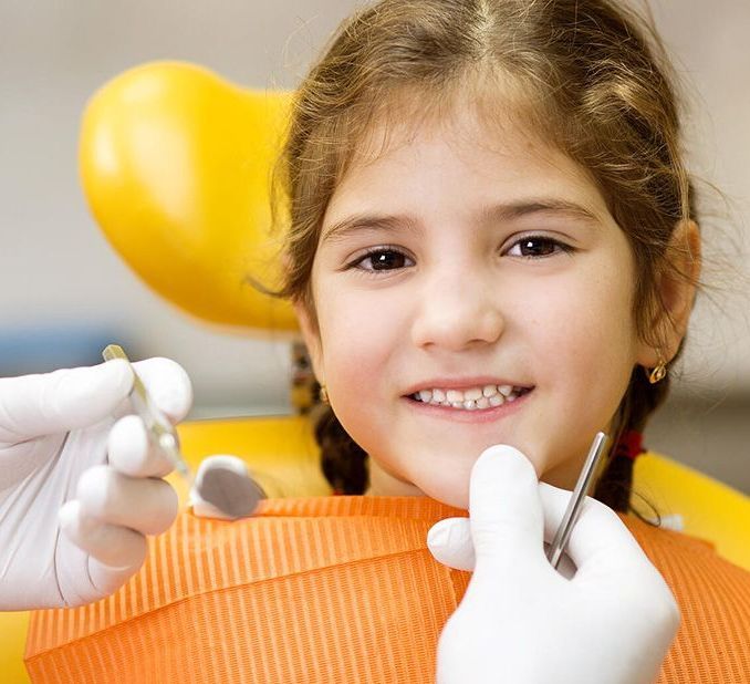 A smiling child sitting in a yellow dental chair with a dentist’s gloved hands holding tools nearby.