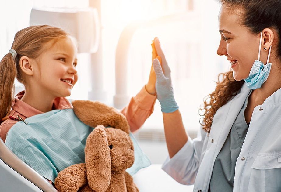 A smiling child holding a stuffed animal high-fives a dentist in a clinical setting.
