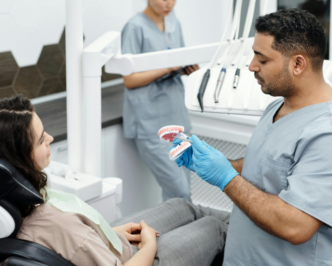 A dentist in blue scrubs and gloves explains a dental model to a patient seated in a chair, while a nurse works nearby.