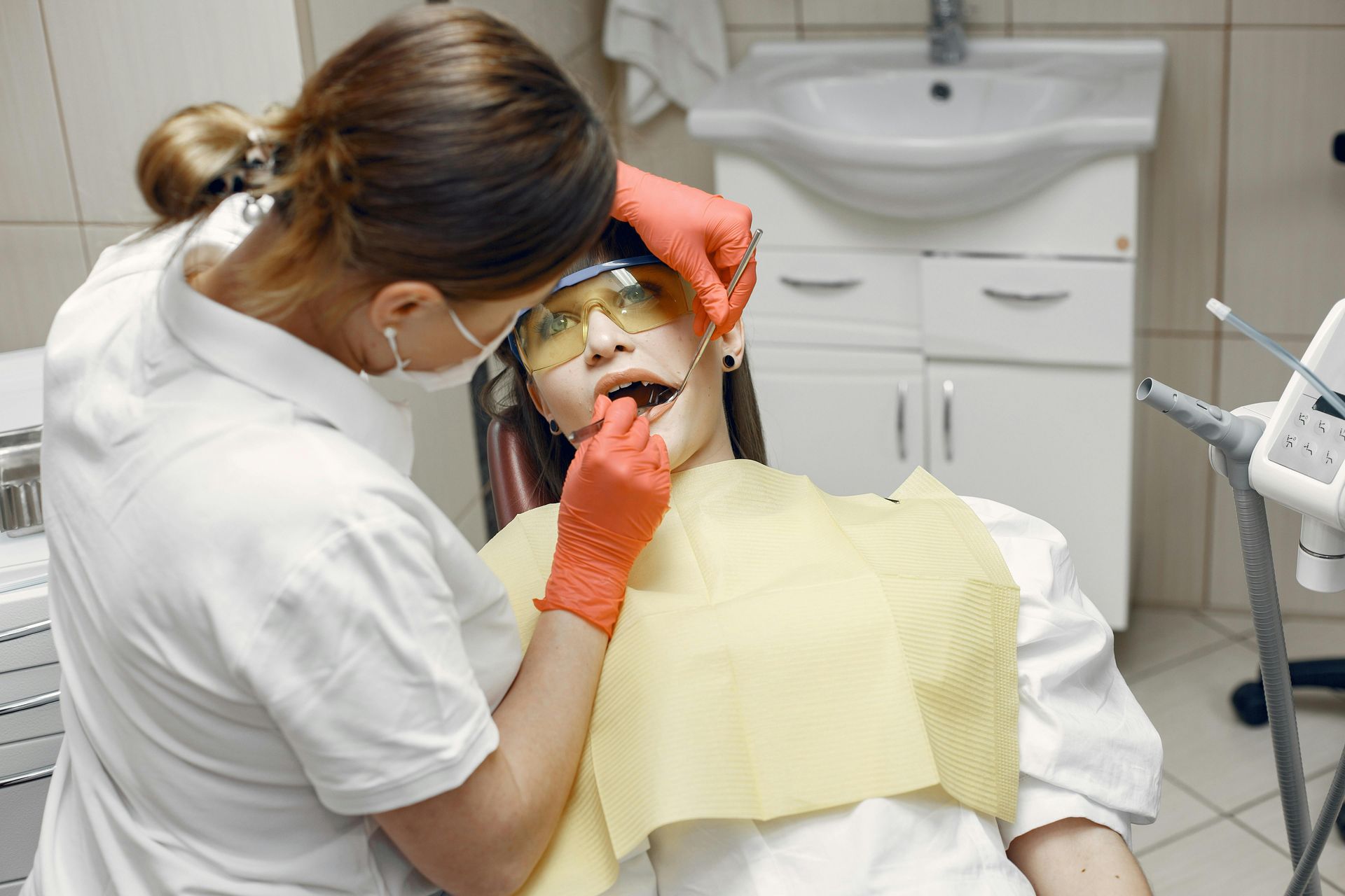 A dental professional wearing orange gloves performs an examination on a patient wearing yellow safety glasses.