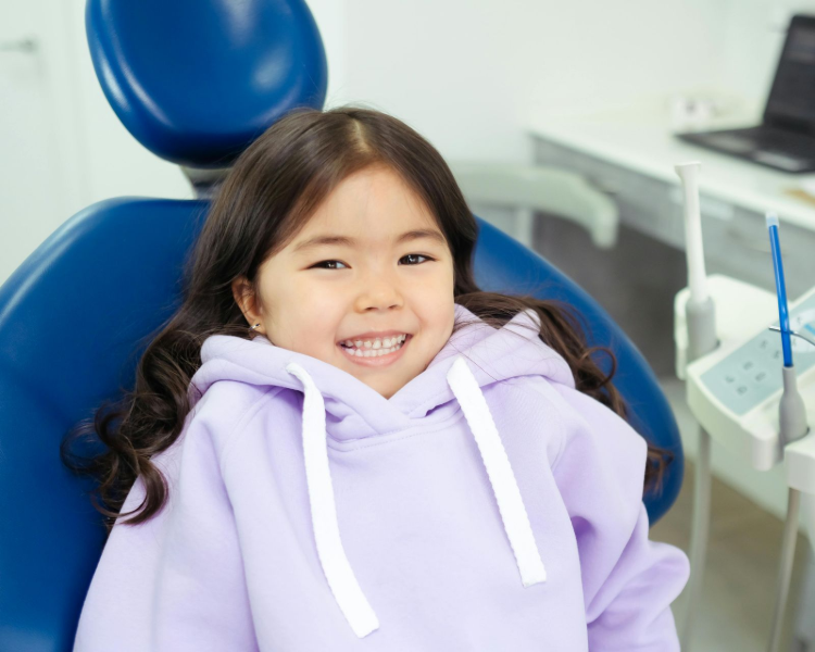 A young child in a lavender hoodie smiling while sitting in a blue dental chair in an exam room.