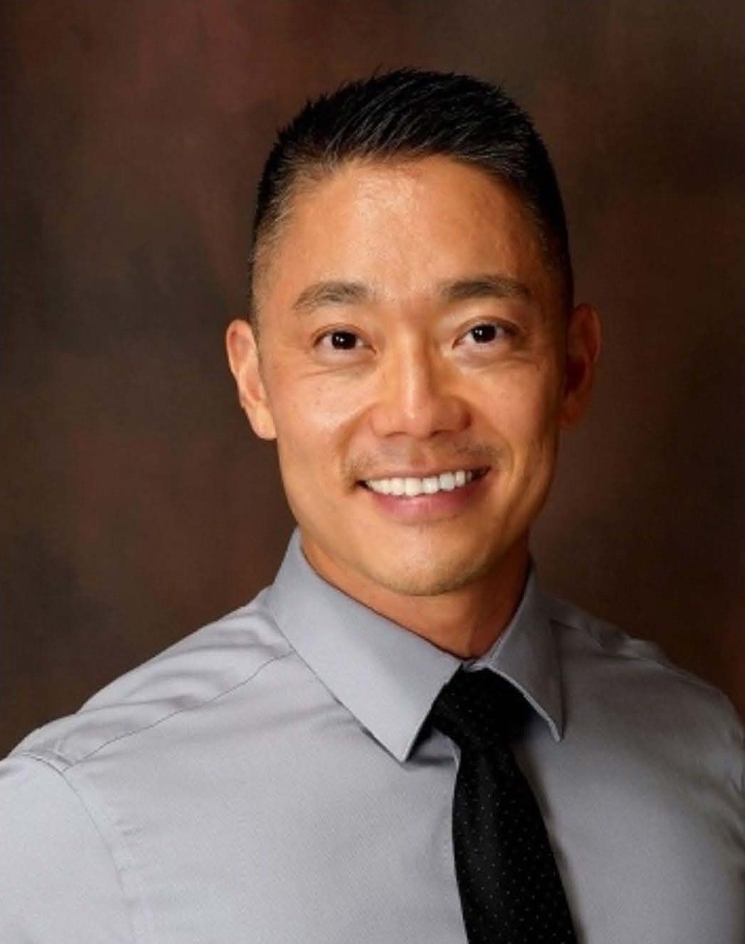A man in a gray shirt and black tie is smiling for the camera.