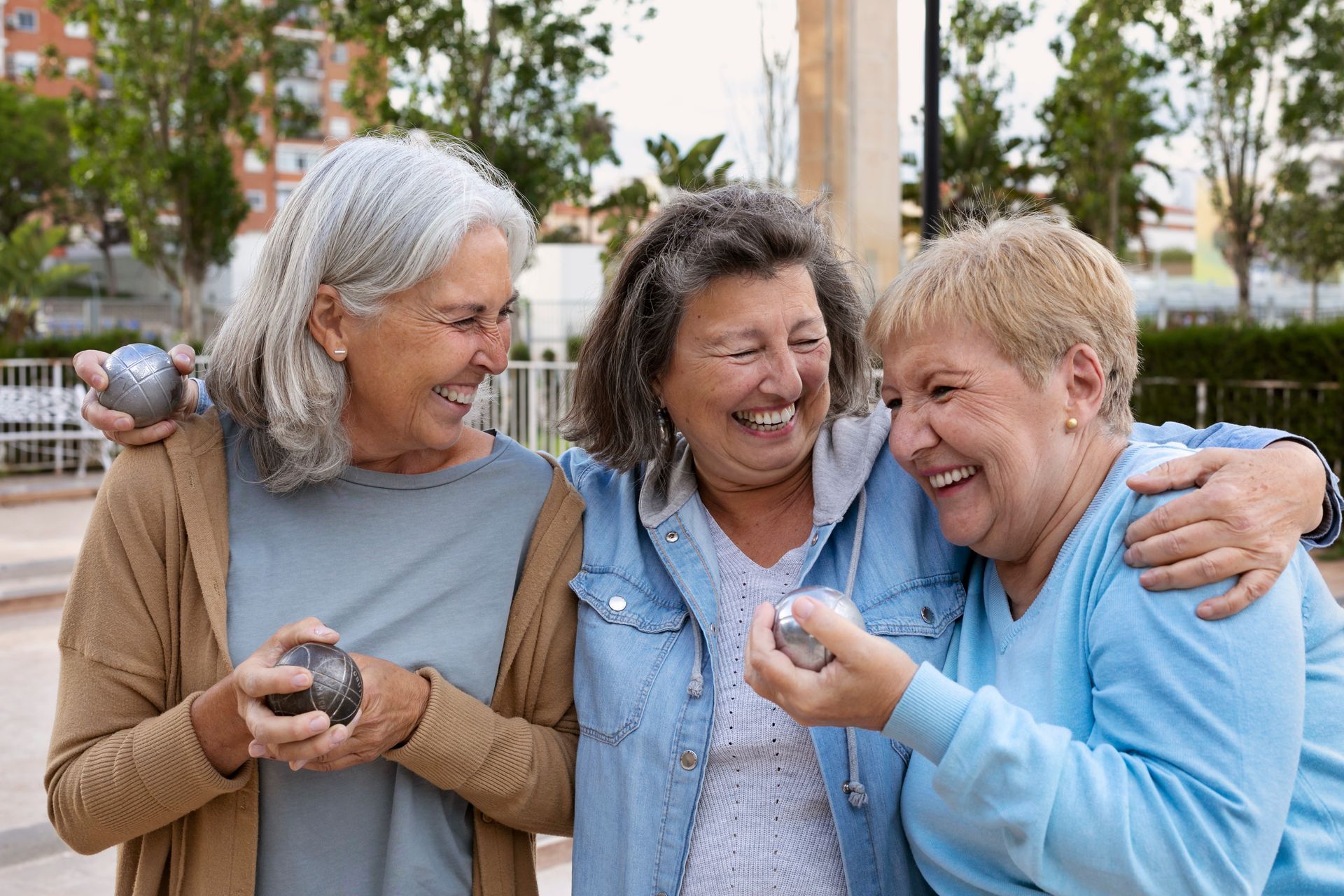Three older women are standing next to each other in a park holding boules and smiling.