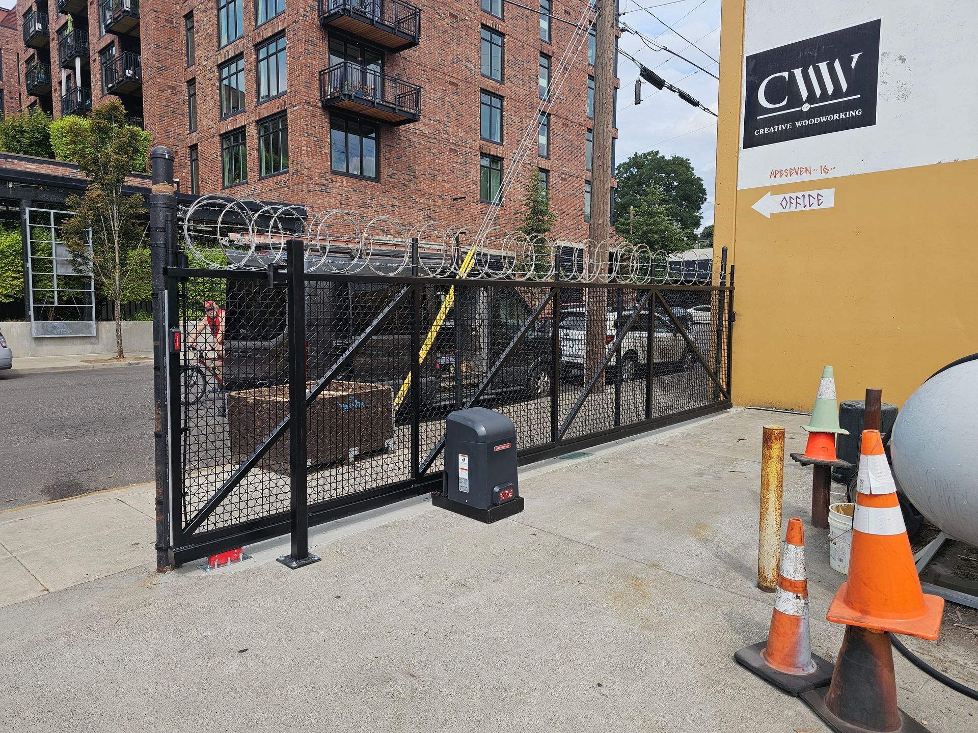 Black security gate with razor wire atop, near brick building and orange traffic cones.