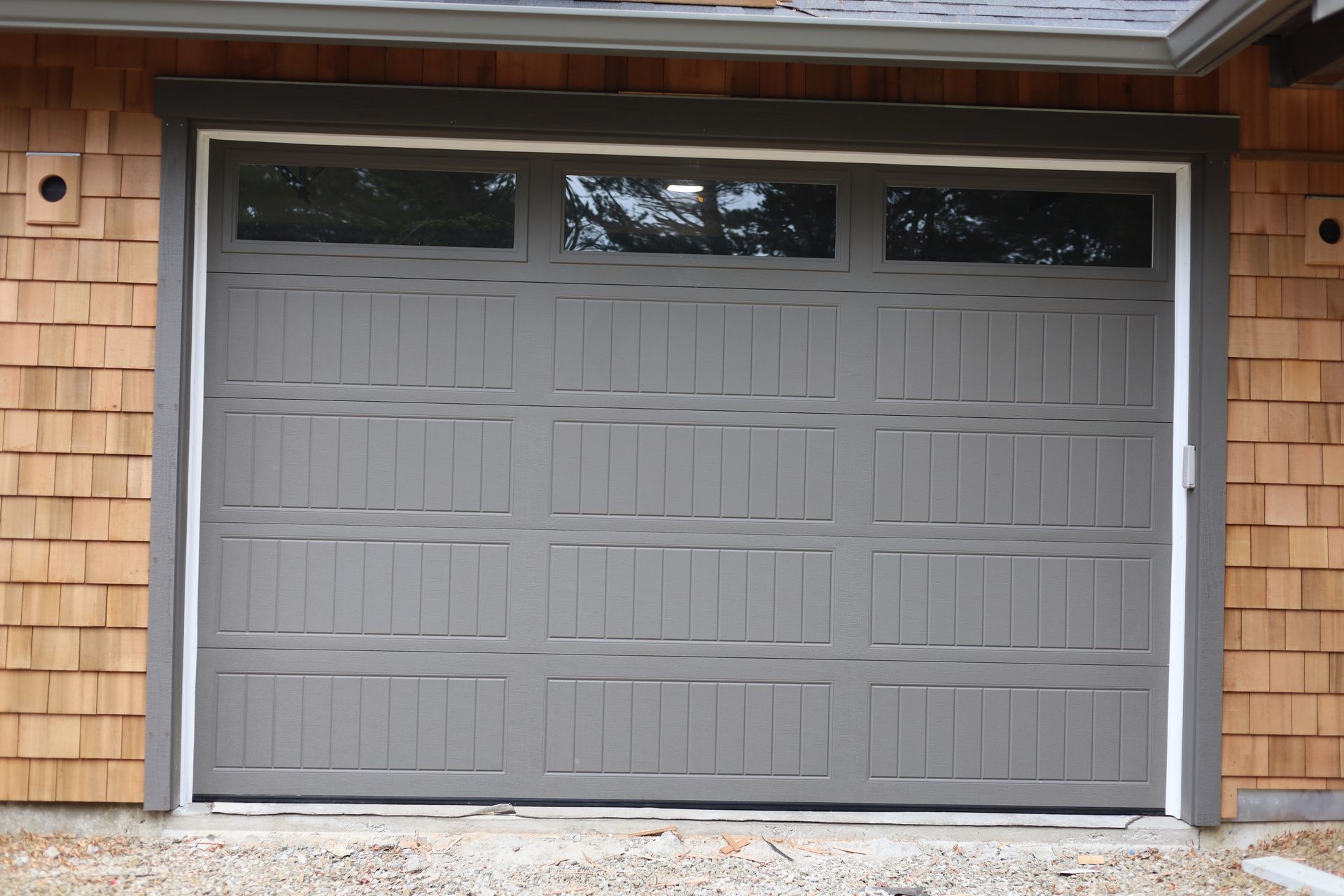 Gray garage door with three small windows above, surrounded by cedar siding.