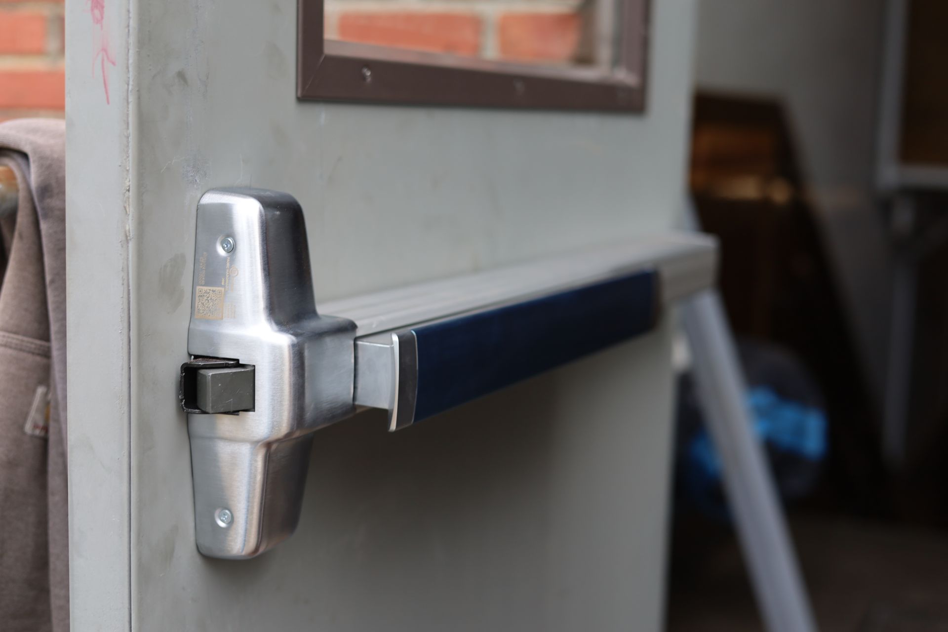 Silver and blue exit bar on a gray door; brick wall visible through the small window.