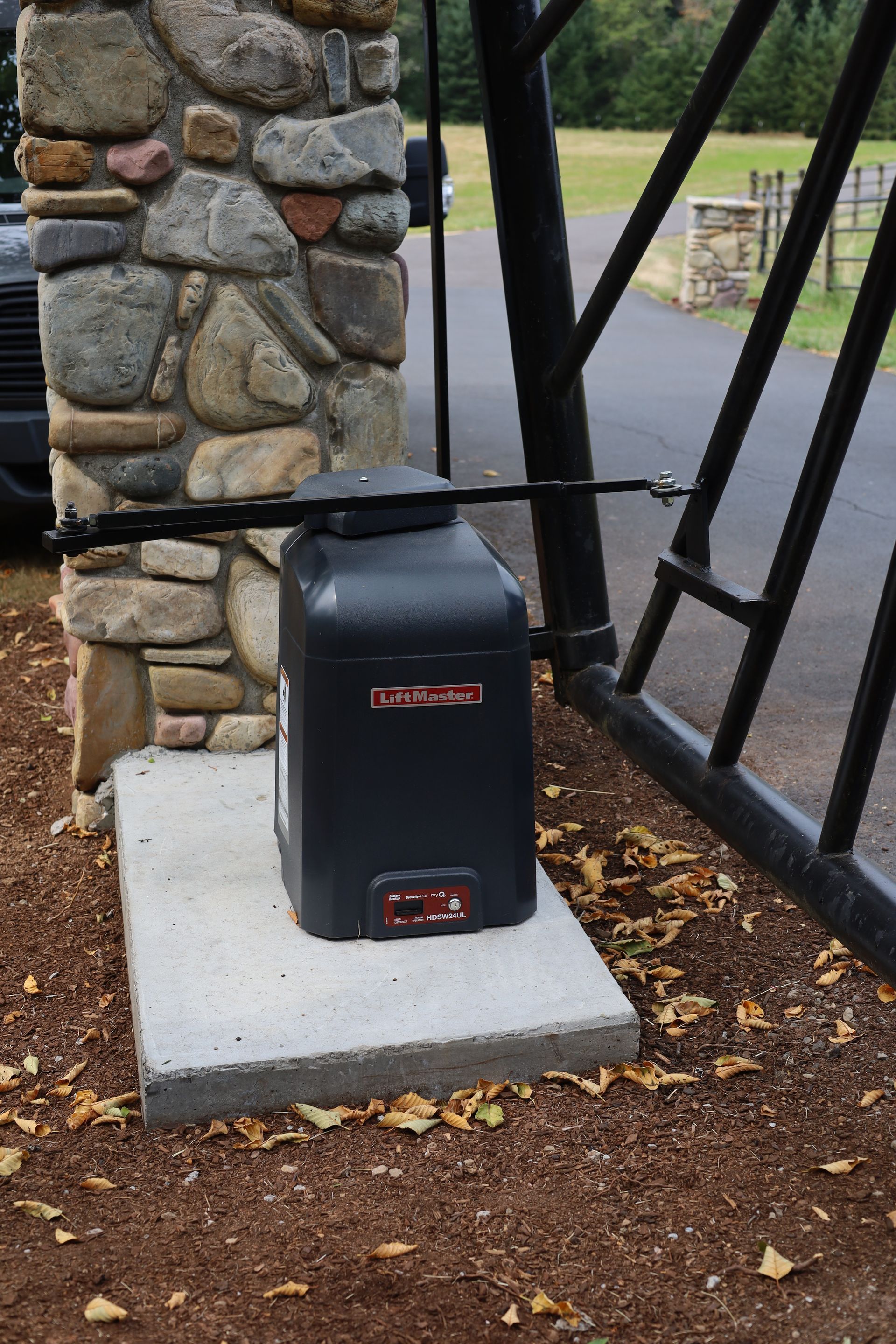 Black gate opener on concrete base next to stone pillar; gate and driveway visible.