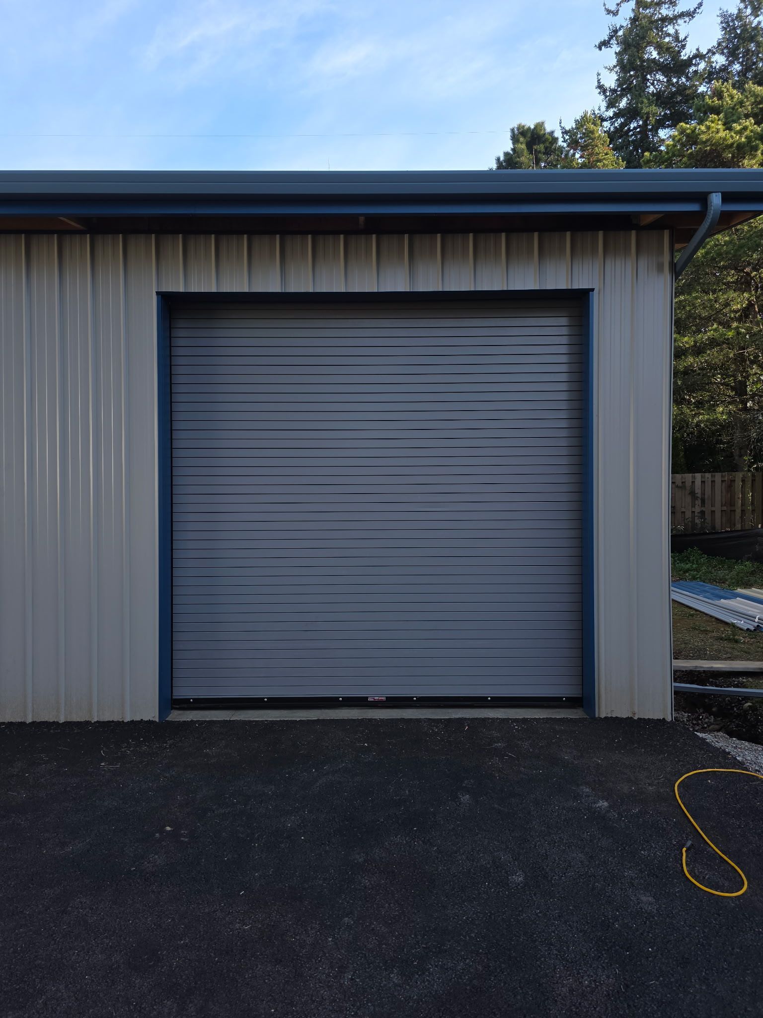 Gray garage door on a tan metal building with a blue trim, set on black asphalt.
