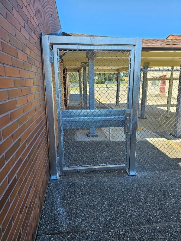 A metal gate secured to a brick wall. The gate has chain link fencing. Outdoors, sunny day.