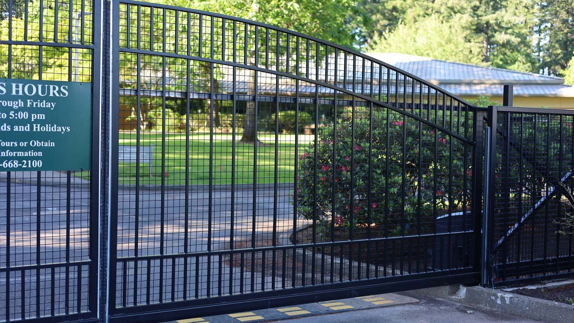 Black metal gate with arched top, leading into a green space with a sign indicating hours.