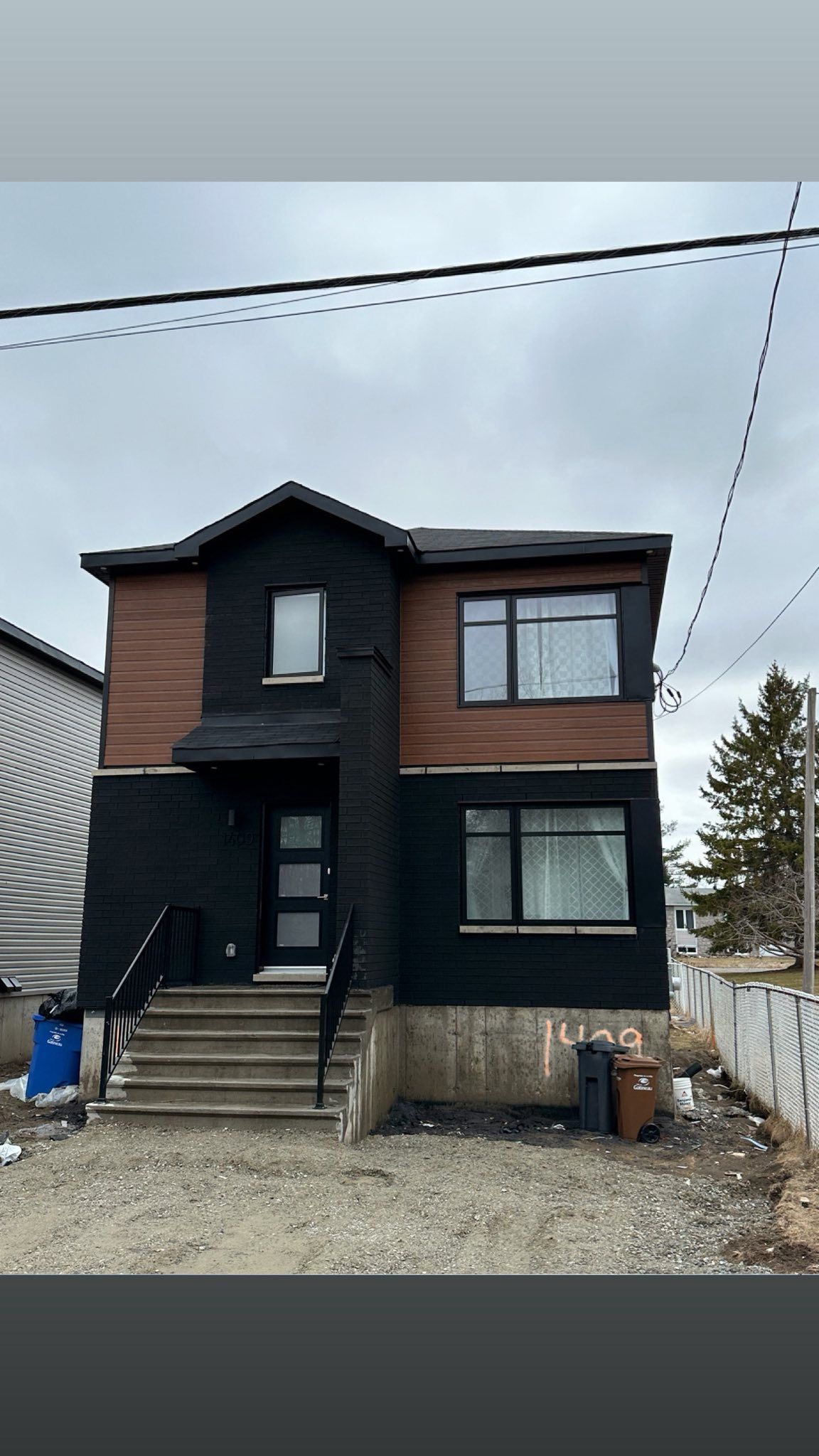 Maison à deux étages avec revêtement noir et bois. Un escalier mène à la porte d'entrée. Ciel gris.
