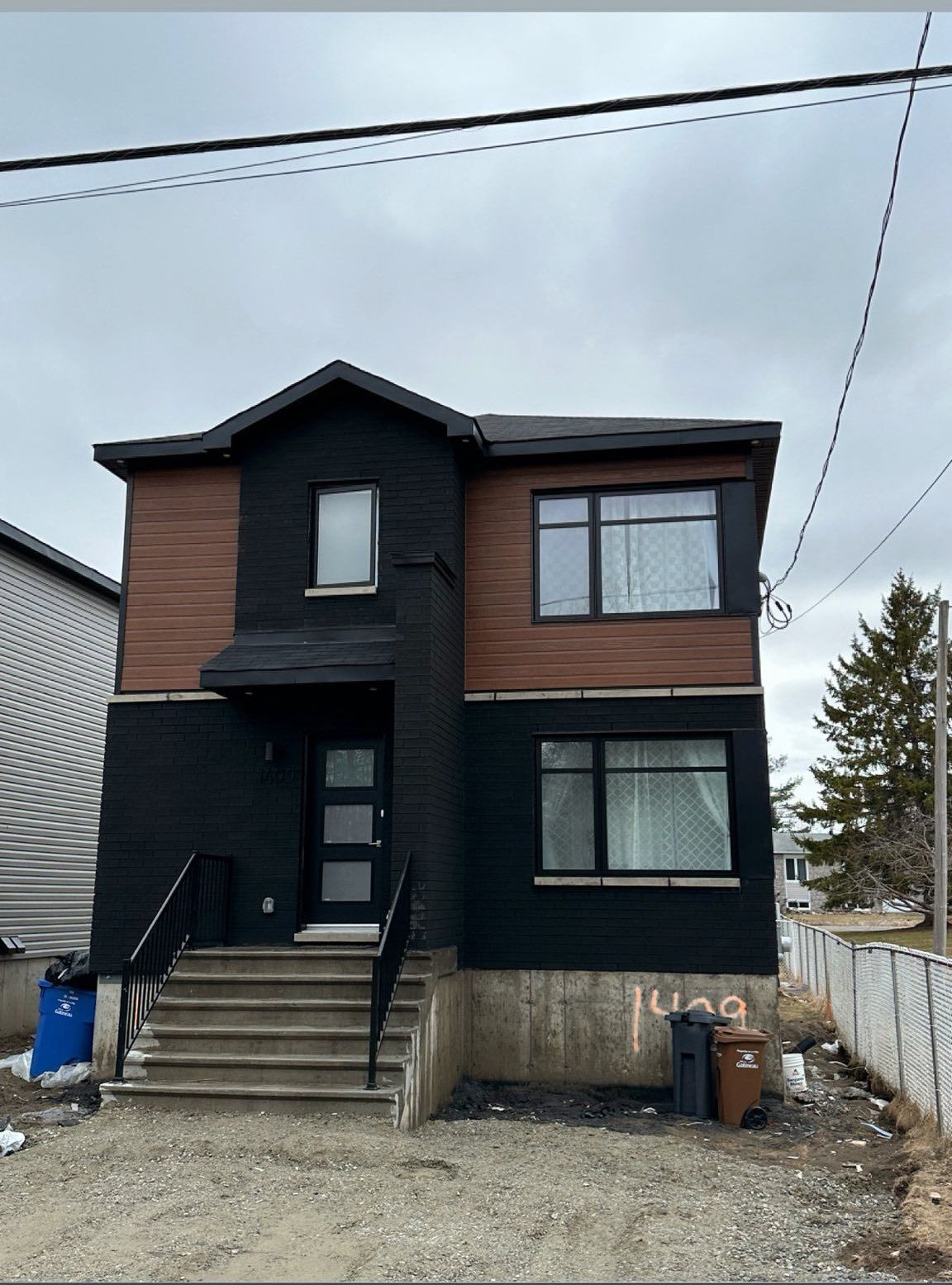 Maison à deux étages avec revêtement noir et touches de marron, escalier gris menant à la porte d'entrée. Journée nuageuse.