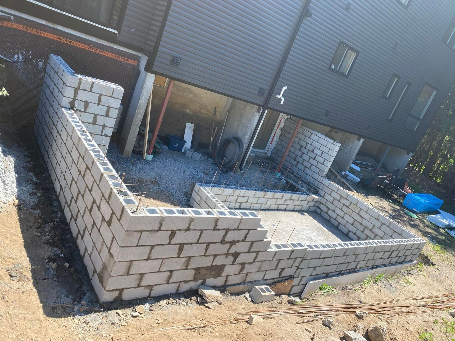 Chantier de construction avec des murs en parpaings en cours de construction, avec une maison en arrière-plan.