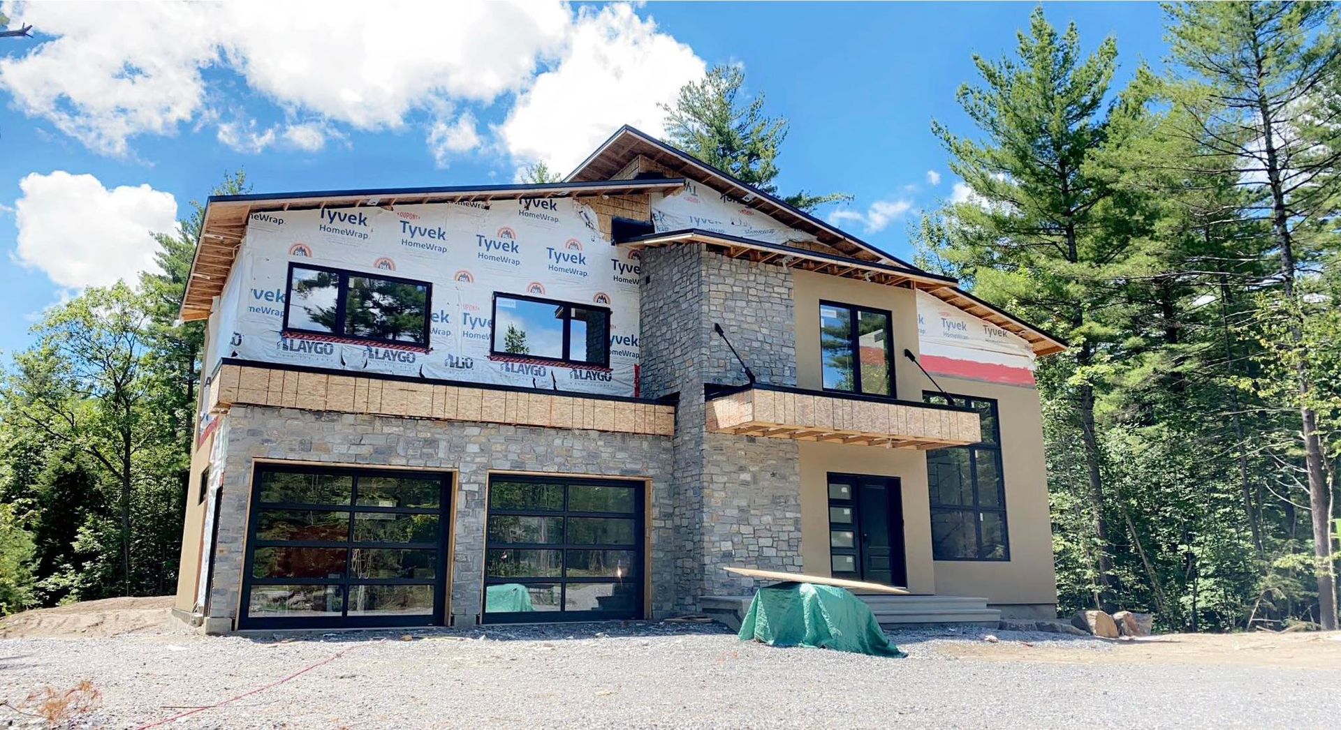 Maison à deux étages en construction avec façade en pierre, grandes fenêtres, portes de garage et arbres en arrière-plan.