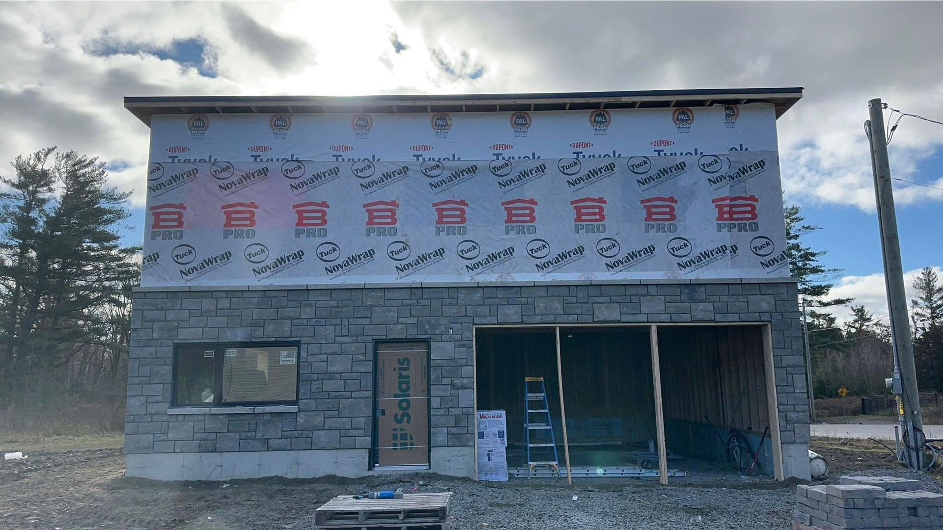 Bâtiment de deux étages en construction; façade en pierre au rez-de-chaussée, revêtement bleu et bois au niveau supérieur, ciel ensoleillé.