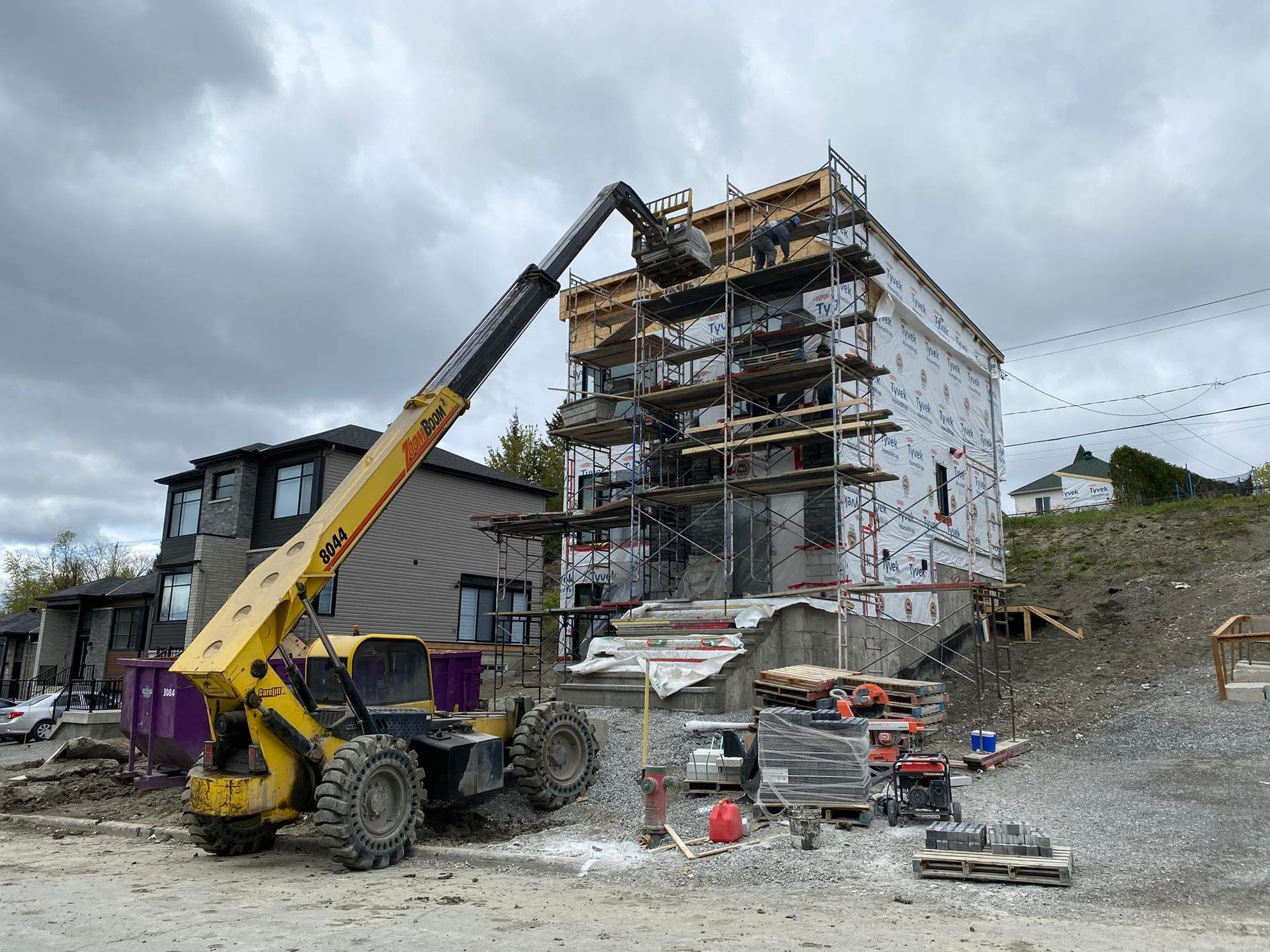 Un chariot télescopique jaune soulève des matériaux vers un immeuble à plusieurs étages en construction, ciel nuageux.