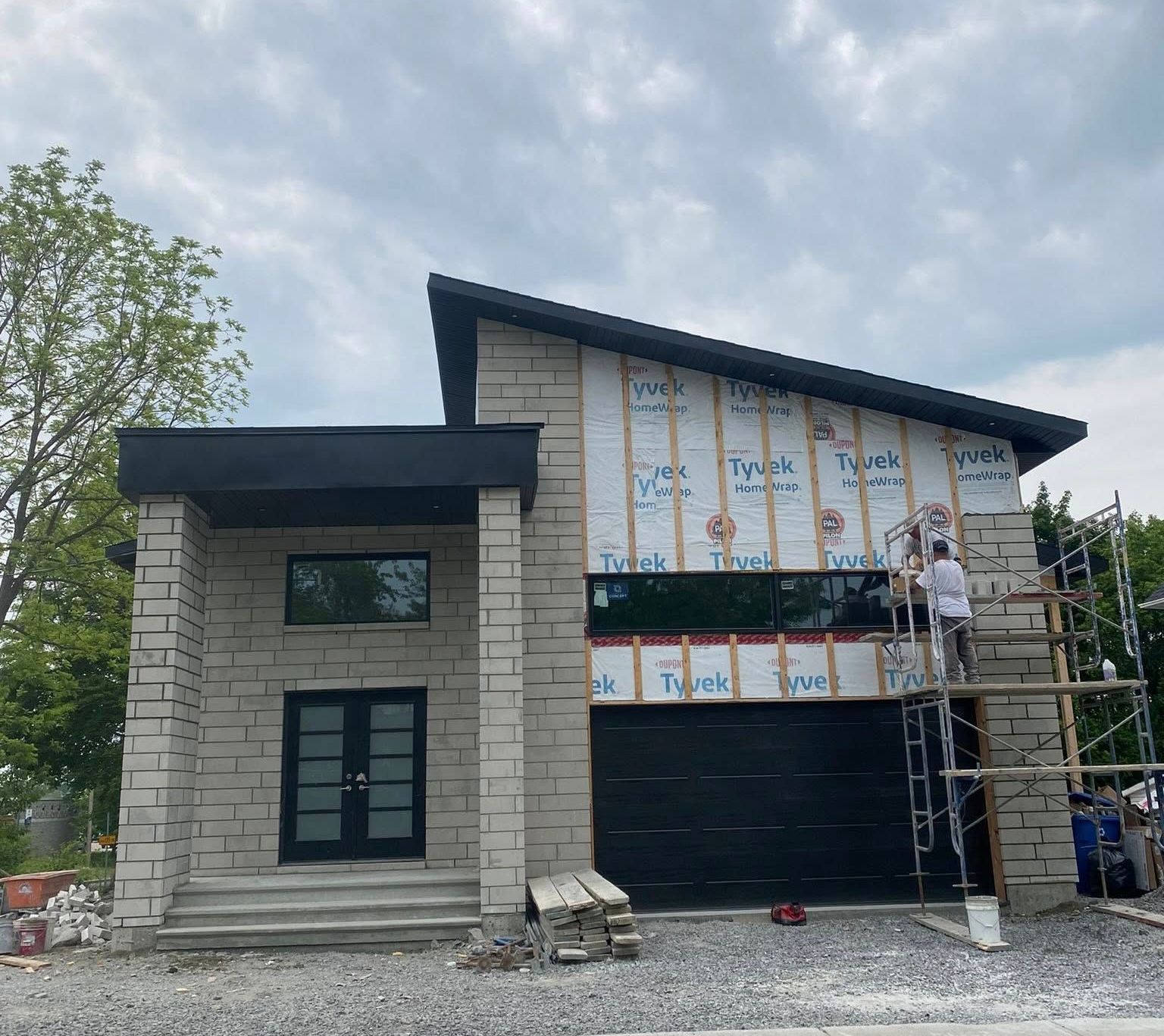 Maison moderne en construction avec façade en pierre, boiseries noires et toit en pente. Ouvriers sur échafaudage.
