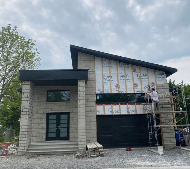 Maison moderne en construction avec façade en pierre, porte de garage noire et ouvriers sur échafaudage.