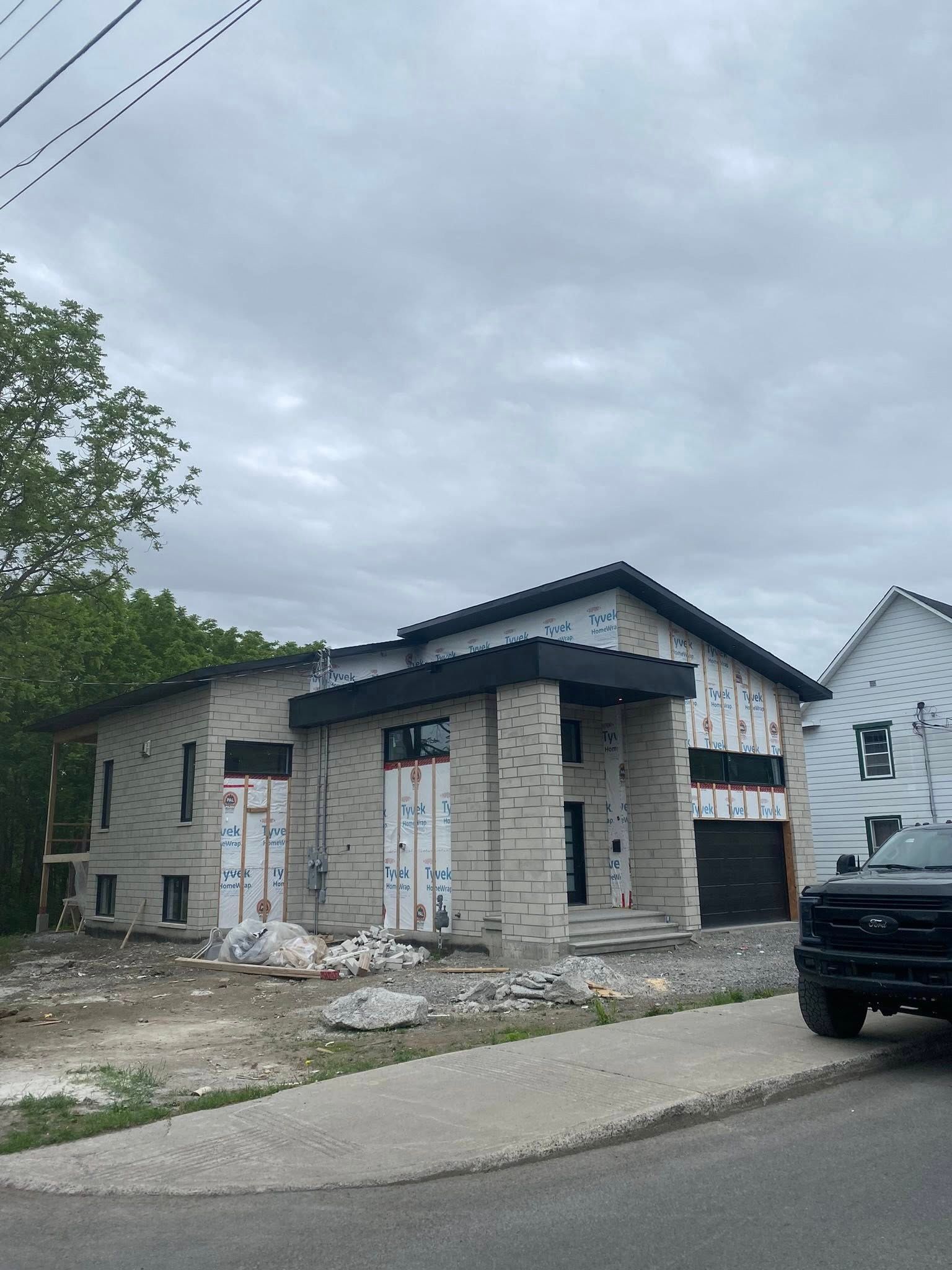 Une maison à deux étages en construction. Brique et bardage gris. Toit et porte de garage foncés. Ciel nuageux.