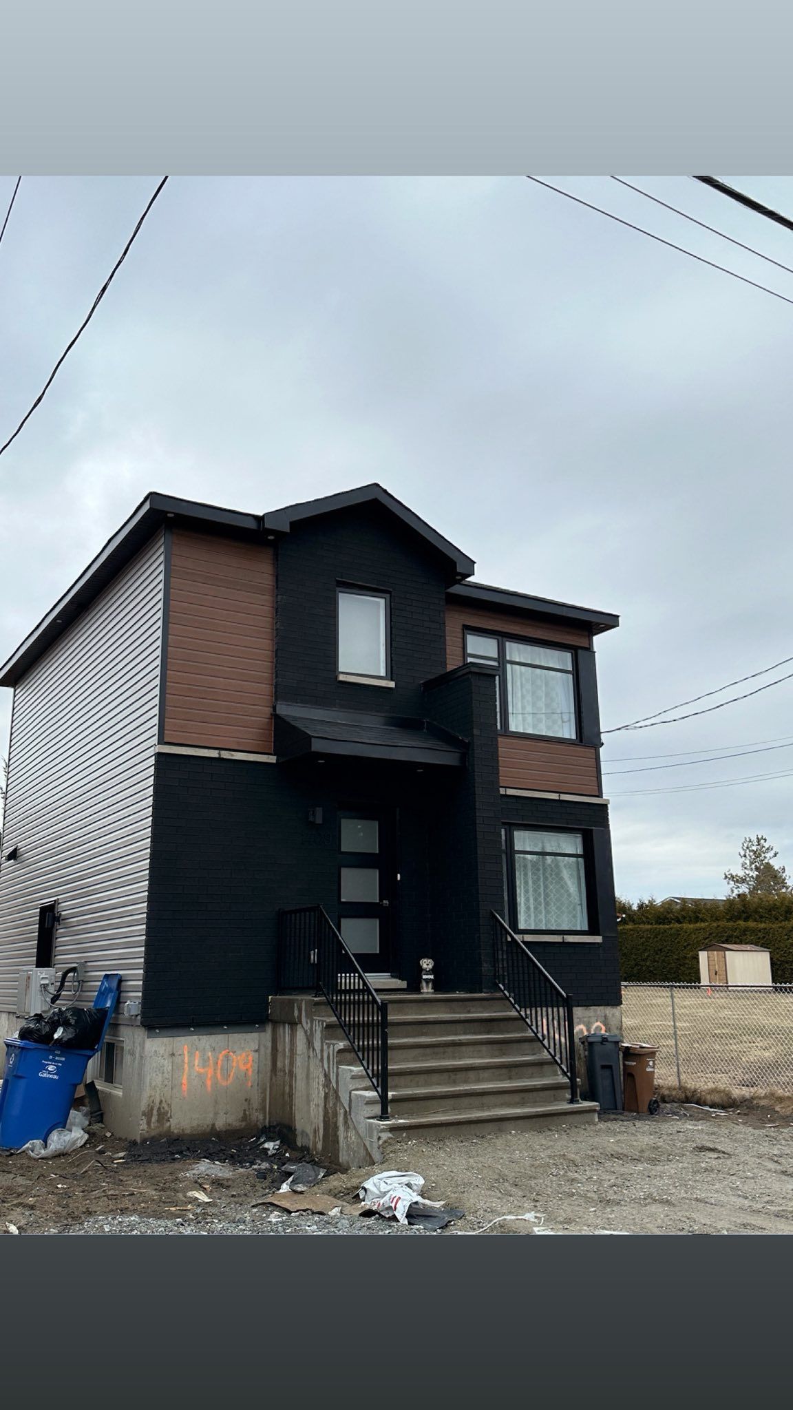Maison à deux étages avec revêtement noir, accent marron et revêtement argenté en spirale à gauche ; ciel gris.