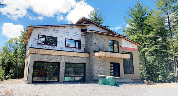 Une maison à deux étages en construction avec extérieur en pierre et stuc, portes de garage en verre et entourée d'arbres.