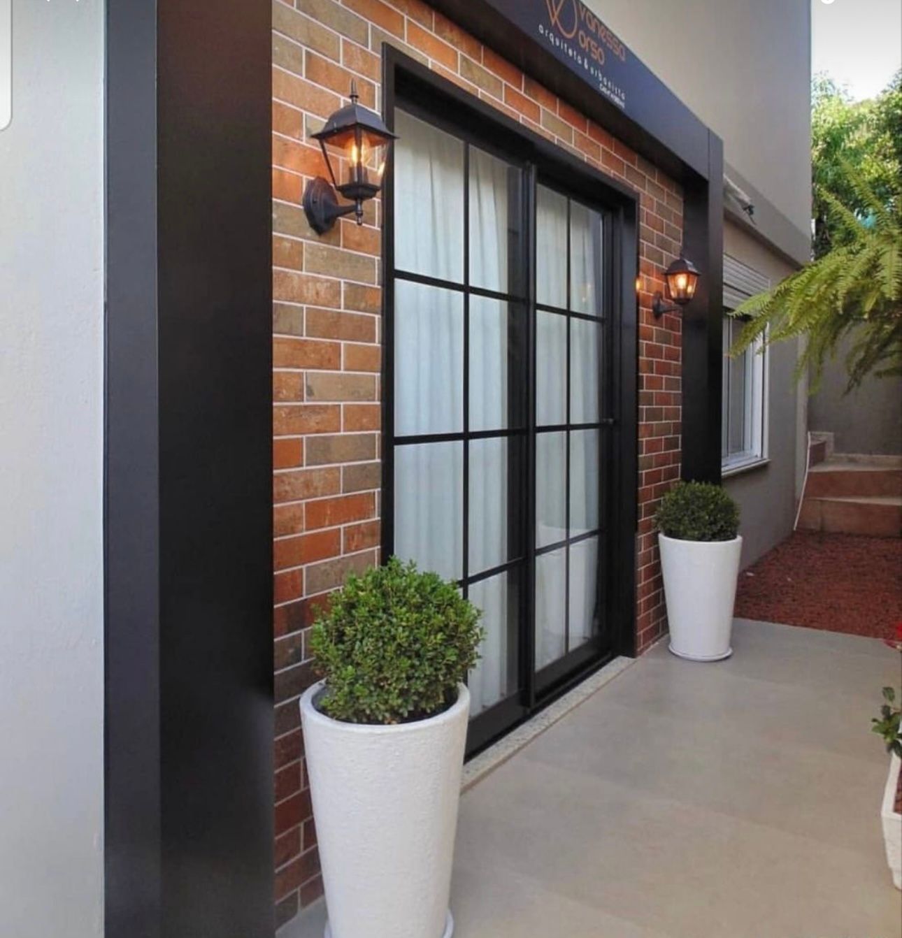 Façade en brique avec porte coulissante à cadre noir, appliques extérieures et plantes en pot.