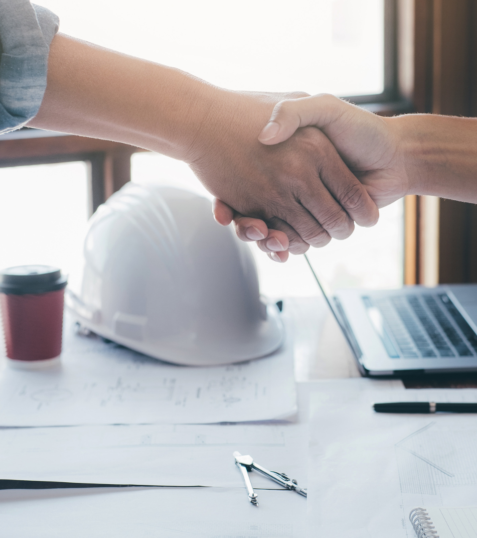 Two people shaking hands over a desk with architectural plans, a hard hat, and a laptop.