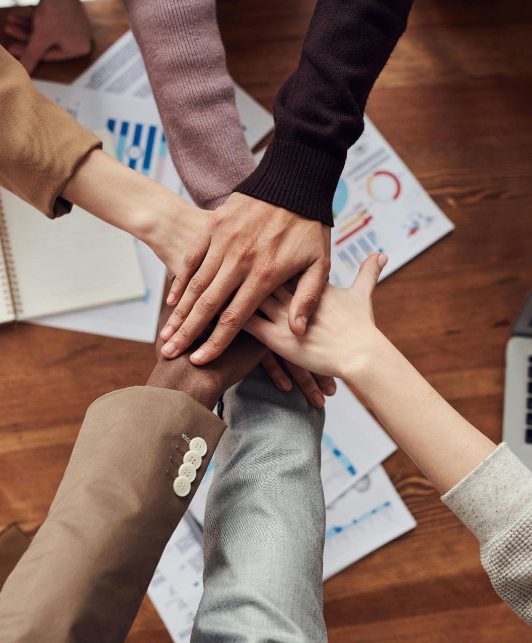 Hands stacked in a team huddle on a wooden table. Papers with graphs, a laptop, and a notebook surround them.