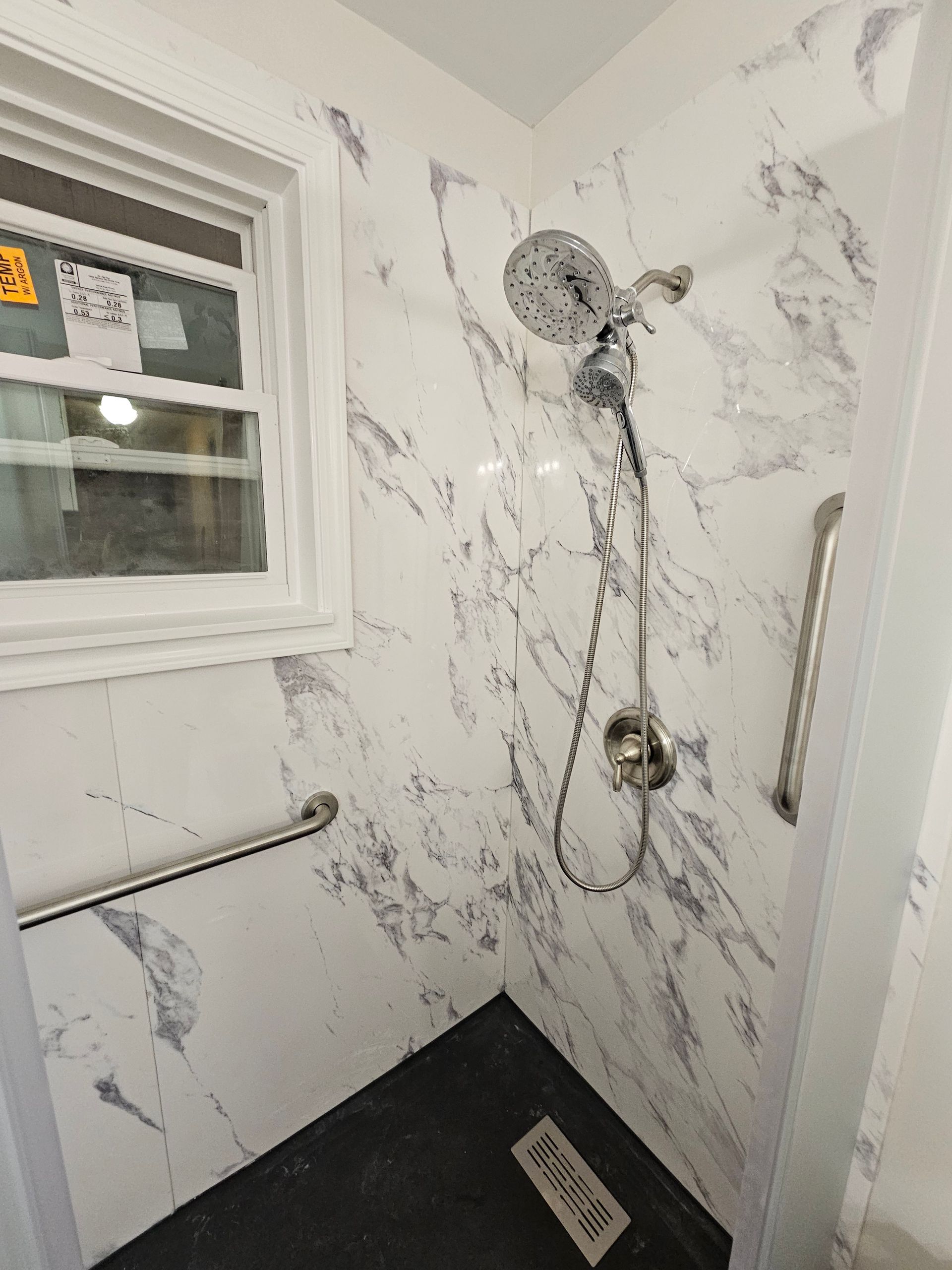 Shower stall with marble-patterned walls and dark floor, featuring a window and grab bars for accessibility.