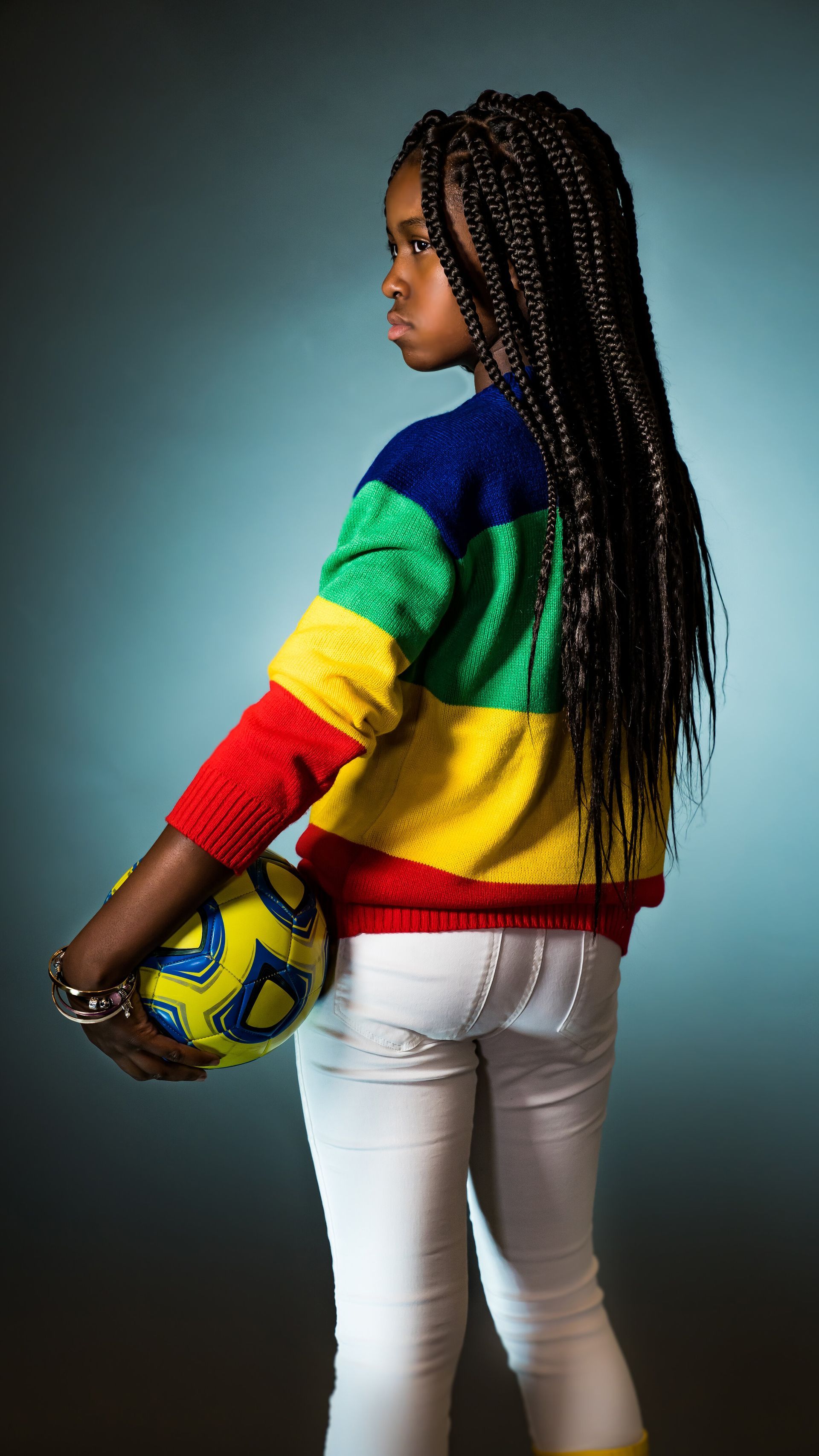A young girl in a rainbow sweater is holding a soccer ball.