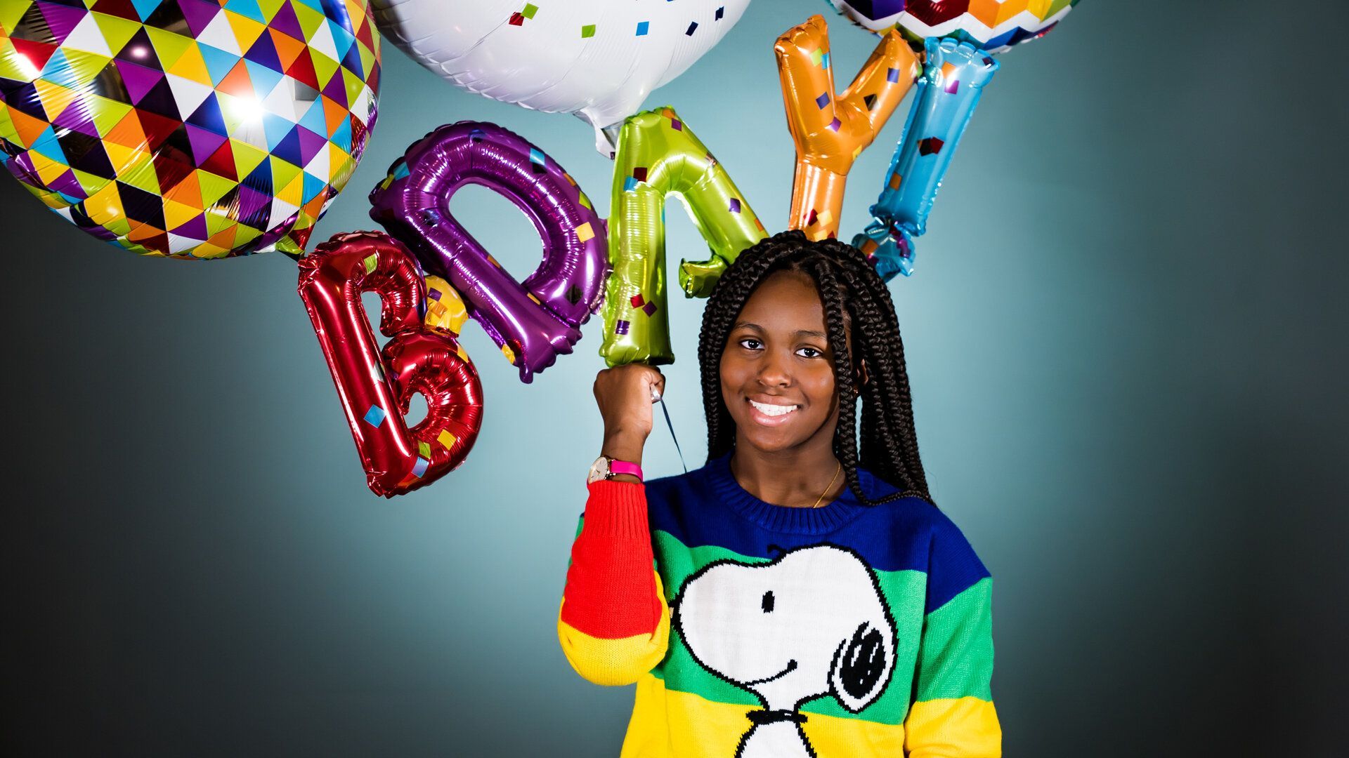 A girl in a snoopy sweater is holding a bunch of birthday balloons.