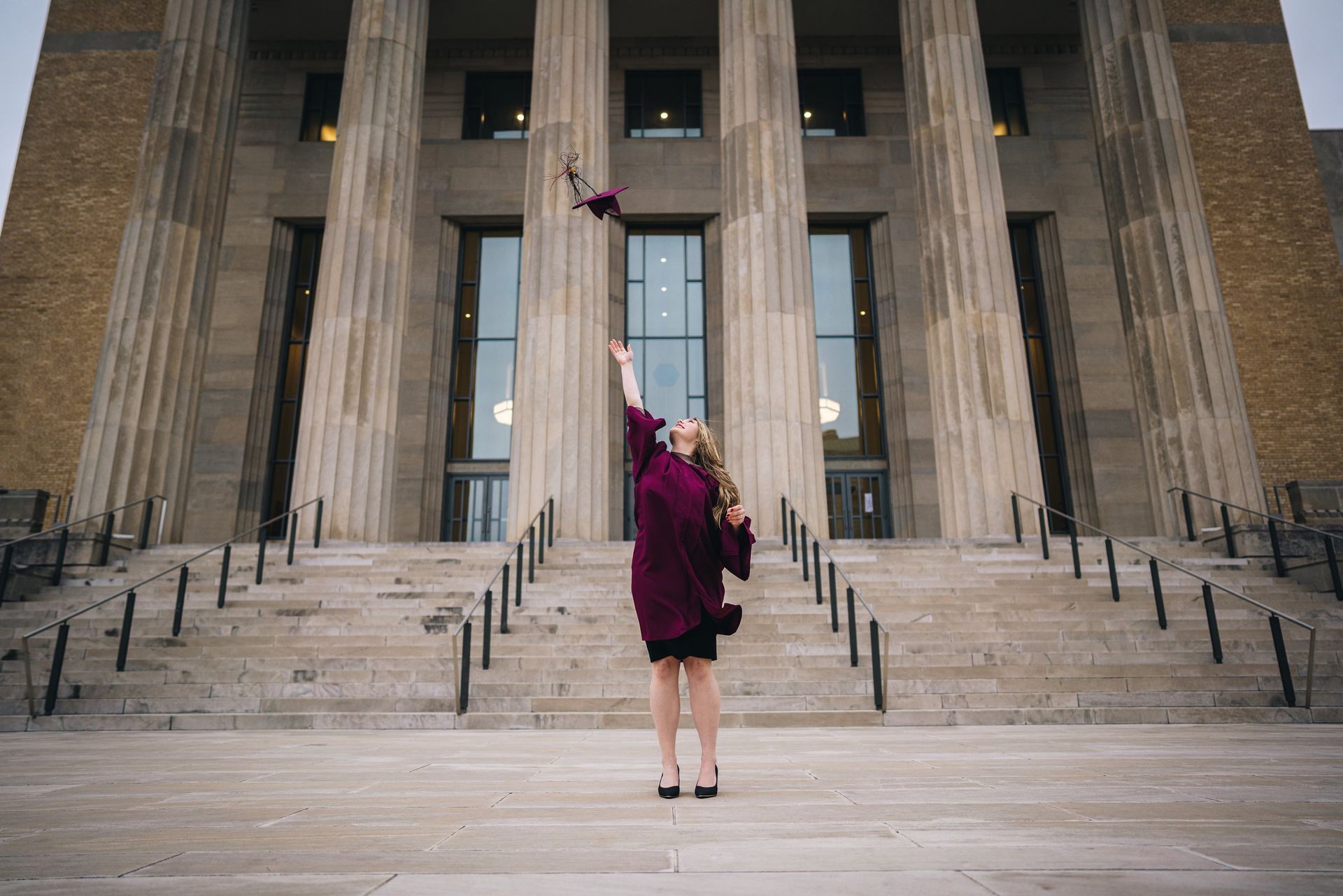 A woman in a graduation cap and gown is throwing her cap in the air in front of a building.